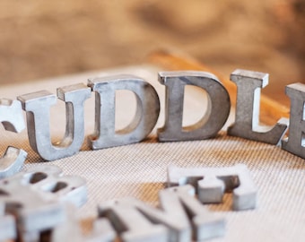 May include: Metal letters spelling out the word "cuddle" in a silver finish. The letters are arranged in a semi-circle on a textured surface.