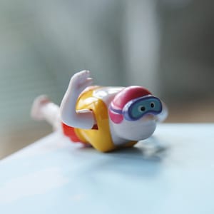 May include: A white plastic toy swimmer with a yellow life vest, pink swim cap, and blue goggles. The toy is in a diving pose with one arm raised.