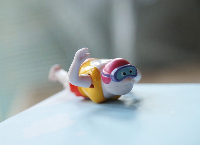 May include: A white plastic toy swimmer with a yellow life vest, pink swim cap, and blue goggles. The toy is in a diving pose with one arm raised.