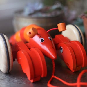 May include: A red wooden toy lobster with black eyes and a pull string. The lobster has orange and yellow stripes on its body and wooden wheels.