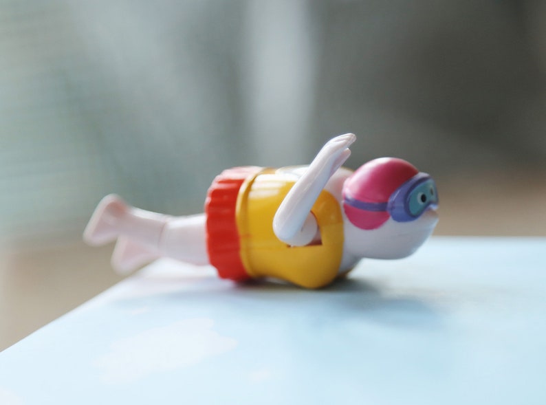 May include: A white plastic wind-up swimmer toy with yellow and red swim trunks, a pink swim cap, and purple goggles. The toy is lying on a blue surface.