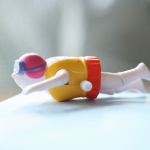 May include: A yellow and red wind-up toy swimmer with a pink and purple diving mask. The swimmer is in a diving position with arms outstretched.