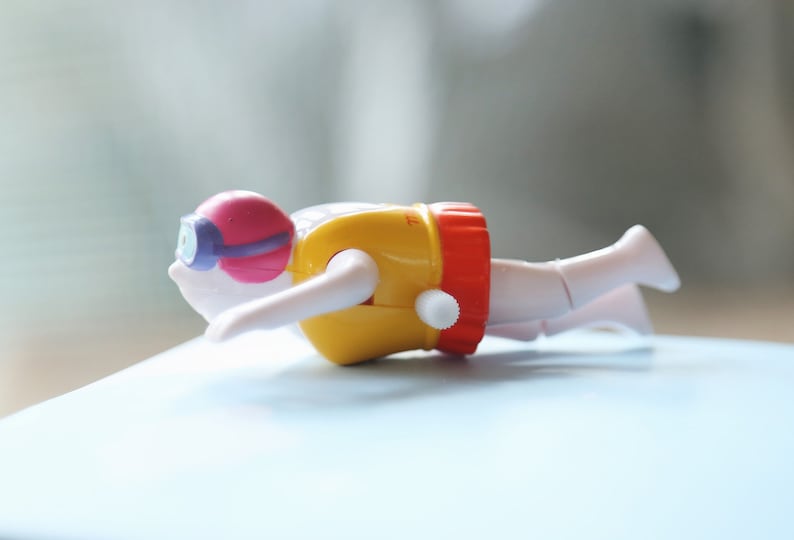 May include: A yellow and red wind-up toy swimmer with a pink and purple diving mask. The swimmer is in a diving position with arms outstretched.