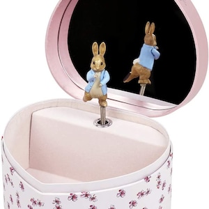 May include: A pink heart-shaped music box with a white interior and a floral pattern. The lid is open and features a rotating figurine of Peter Rabbit.
