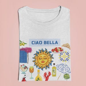 May include: A white t-shirt with a colorful illustration of Italian-themed imagery, including a sun, a lemon, a lobster, a glass of wine, a bottle of olive oil, a beach umbrella, and a blue door. The text "Ciao Bella" is printed in blue on a white rectangle.