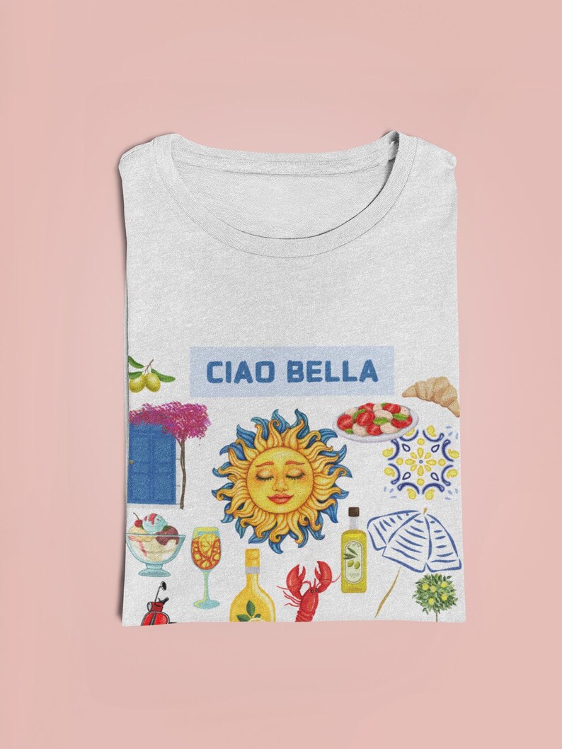 May include: A white t-shirt with a colorful illustration of Italian-themed imagery, including a sun, a lemon, a lobster, a glass of wine, a bottle of olive oil, a beach umbrella, and a blue door. The text "Ciao Bella" is printed in blue on a white rectangle.