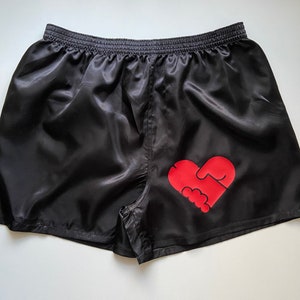 Pode incluir: Shorts de cetim preto com um gr&aacute;fico vermelho em forma de cora&ccedil;&atilde;o que apresenta duas m&atilde;os dadas.