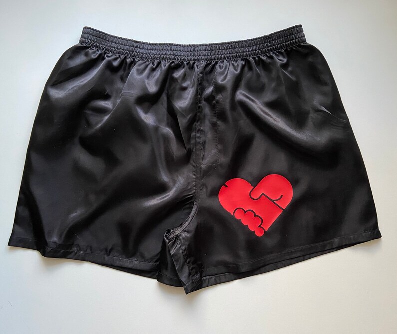 Pode incluir: Shorts de cetim preto com um gr&aacute;fico vermelho em forma de cora&ccedil;&atilde;o que apresenta duas m&atilde;os dadas.
