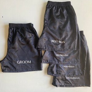 Pode incluir: Cinco cal&ccedil;&otilde;es de desporto de nylon preto com texto branco. Os cal&ccedil;&otilde;es est&atilde;o empilhados uns sobre os outros. O cal&ccedil;&atilde;o de cima diz "Best Man". Os tr&ecirc;s seguintes dizem "Groomsman". O cal&ccedil;&atilde;o de baixo diz "Groom".