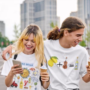 May include: Two people wearing white t-shirts with graphic designs. The person on the left is holding a phone and smiling. The person on the right is holding an ice cream cone and smiling. The t-shirt on the left has a variety of colorful illustrations, including a sun, a lobster, a bottle of wine, and a scooter. The t-shirt on the right has a lemon, a plus sign, a violin, an equals sign, and a bottle of liquor.
