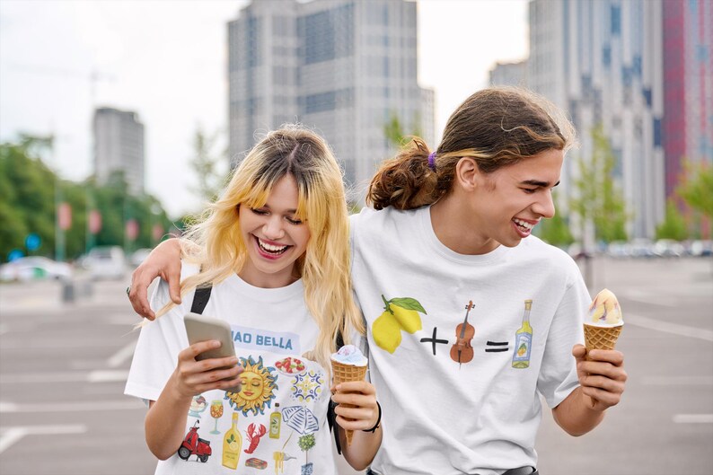 May include: Two people wearing white t-shirts with graphic designs. The person on the left is holding a phone and smiling. The person on the right is holding an ice cream cone and smiling. The t-shirt on the left has a variety of colorful illustrations, including a sun, a lobster, a bottle of wine, and a scooter. The t-shirt on the right has a lemon, a plus sign, a violin, an equals sign, and a bottle of liquor.