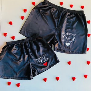 Pode incluir: Dois pares de cal&ccedil;&otilde;es de cetim preto com texto branco. Um par diz "PROPERTY OF Cheryl EST. 2021" e o outro diz "PROPERTY OF Nisha".