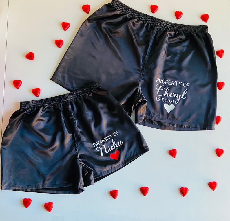 Pode incluir: Dois pares de cal&ccedil;&otilde;es de cetim preto com texto branco. Um par diz "PROPERTY OF Cheryl EST. 2021" e o outro diz "PROPERTY OF Nisha".