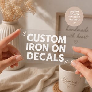 May include: A rectangular, transparent sheet featuring the white text CUSTOM IRON ON DECALS in a bold, sans-serif font. The sheet includes small decorative white line accents around the text. The design is presented on a clear, flexible material.