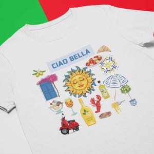 May include: White t-shirt with a colorful graphic design featuring the text "Ciao Bella" and various Italian-themed illustrations, including a sun, a scooter, a bottle of limoncello, and a lemon.