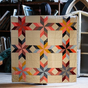 Puede incluir: Una colcha con un diseño de patchwork que presenta un patrón repetitivo de estrellas en tonos de rosa, rojo, naranja, azul y negro. La colcha está hecha de una variedad de telas y se muestra sobre un fondo de ventanas antiguas.