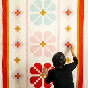May include: A white quilt with a red, pink, and blue floral pattern. The quilt is made up of squares with a geometric design. A person is holding the quilt up.