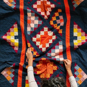 Könnte beinhalten: Eine Person hält eine farbenfrohe Patchwork-Decke mit dunkelblauem Hintergrund hoch. Die Decke besteht aus Quadraten in verschiedenen Farben und Mustern, darunter Rot, Orange, Gelb, Grün, Blau und Lila. Die Decke ist in der Mitte gefaltet, und die Person hält die Ränder der Decke hoch.