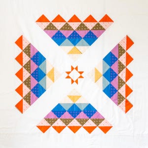 May include: A white quilt with a geometric pattern of orange, pink, blue and yellow triangles. The quilt is made up of smaller squares that are sewn together to create the larger design.