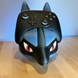 Könnte beinhalten: Schwarzer und blauer Nintendo Switch Controller-Halter im Lucario-Design. Der Controller ist schwarz mit weißen Knöpfen und einem Steuerkreuz. Der Halter hat rotäugige Lucario-Merkmale und ist zur Aufnahme des Controllers konzipiert.