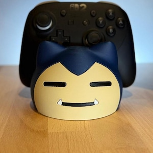 Könnte beinhalten: Ein dunkelblauer und cremefarbener Controller-Ständer in Charakterform mit einem stilisierten Gesicht mit geschlossenen Augen und zwei kleinen weißen Fangzähnen, platziert vor einem schwarzen kabellosen Gaming-Controller mit Standardtasten.