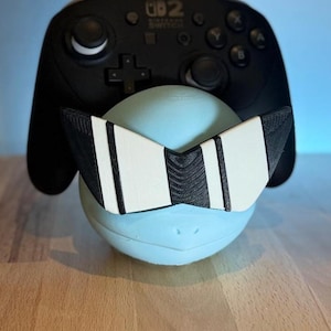 Può includere: Una sfera azzurra con occhiali da sole bianchi e neri davanti a un controller Nintendo Switch nero. Gli occhiali da sole hanno un design geometrico con strisce bianche. Il controller è nero.