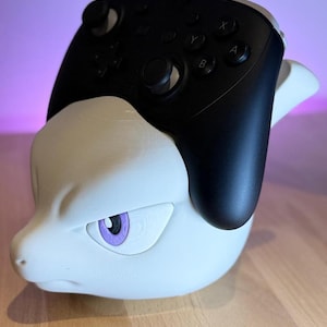 Può includere: Un supporto per controller bianco stampato in 3D a forma di testa di personaggio dei cartoni animati, con un occhio viola. Un controller nero con pulsanti e joystick è appoggiato sopra.