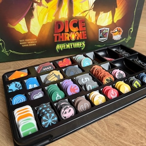 Peut inclure: Jeu de société "Dice Throne Adventures" avec un insert en plastique noir rempli de pièces colorées. La boîte de jeu présente des illustrations de personnages et le titre du jeu. Les pièces du jeu sont de formes et de couleurs variées.