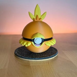 Peut inclure: Une figurine orange et jaune imprimée en 3D représentant un personnage de dessin animé avec un grand œil et une crête en forme de flamme. La figurine a un corps rond, des accents noirs et repose sur un dessous de verre gris foncé.
