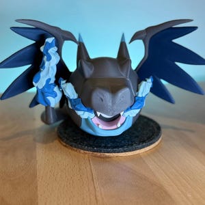 Può includere: Figura stampata in 3D di una creatura simile a un drago, con corpo marrone, ali blu e fiamme blu e bianche. La figura è posta su una base rotonda grigio scuro.