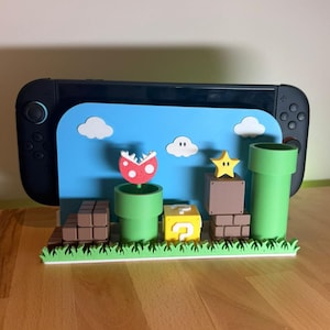 Peut inclure: Une console Nintendo Switch noire est présentée avec un support personnalisé représentant des éléments du jeu Super Mario. Le support comprend des tuyaux verts, des blocs bruns, un bloc jaune avec un point d'interrogation, une étoile, une plante Piranha et des nuages blancs sur fond bleu.
