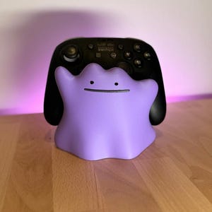 Op de afbeelding: Een paarse Ditto-vormige controllerstandaard met een zwarte Nintendo Switch Pro-controller. De standaard heeft een eenvoudig, lachend gezicht. De controller heeft knoppen en joysticks.