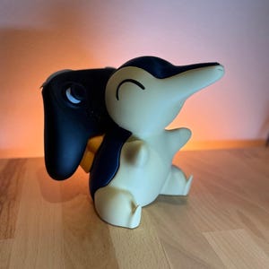 Könnte beinhalten: Ein Cyndaquil-förmiger Controller-Halter in Creme, Marineblau und Gelb. Der Halter ist so konzipiert, dass er einen schwarzen Gamecontroller aufnehmen kann. Die Figur hat eine glatte Oberfläche und steht auf einer Holzoberfläche.