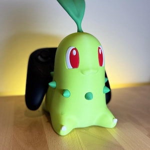 Könnte beinhalten: Ein hellgrüner Chikorita-Controllerhalter mit roten Augen, grünen Stacheln und einem Blatt auf dem Kopf. Ein schwarzer Controller befindet sich in der Halterung. Der Halter steht auf einer Holzoberfläche.