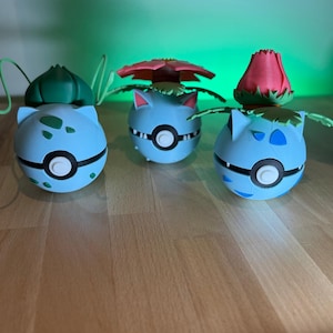 Könnte beinhalten: Drei blaue und grüne Pokémon-Figuren mit verschiedenen farbigen Blüten-artigen Oberteilen. Die Figuren stehen auf einer Holzoberfläche.