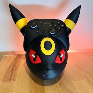 Könnte beinhalten: Schwarzer Nintendo Switch Pro Controller mit individuellem Umbreon-Design. Der Controller hat gelbe Akzente, rote Augen und einen gelben Ring. Ein einzigartiges Zubehör für Gamer.
