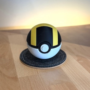 Op de afbeelding: Een bolvormig speelgoed met een zwart-gele bovenhelft en een witte onderhelft. Een ronde witte knop is in het midden aan de voorzijde geplaatst, waar de twee helften samenkomen. Het object is ontworpen met een gladde afwerking en kleurvlakken.