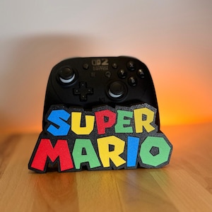 Peut inclure: Un support de manette de jeu vidéo noir avec le texte SUPER MARIO en lettres capitales multicolores en relief. Les lettres sont bleues, rouges, vertes et jaunes sur une base noire. Le support est conçu pour maintenir une manette noire.