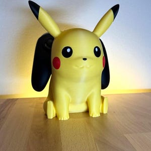 Può includere: Una statuetta gialla di Pikachu con orecchie dalle punte nere e guance circolari rosse. Il personaggio è raffigurato in posizione seduta con due grandi forme nere, solide e arrotondate, che sporgono da dietro la testa.