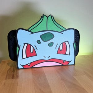 Könnte beinhalten: Ein hellblau-grüner Nintendo Switch-Dock im Bulbasaur-Design. Das Dock zeigt ein Cartoon-Bulbasaur-Gesicht mit roten Augen, einem rosa Mund und grünen Akzenten. Das Dock ist für eine Nintendo Switch-Konsole und Controller konzipiert.
