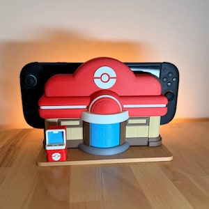 Peut inclure: Modèle imprimé en 3D rouge et blanc d'un bâtiment avec un logo circulaire sur le toit, ressemblant à un Centre Pokémon. Le modèle a une porte bleue et un petit kiosque rouge. Une Nintendo Switch noire est derrière le modèle.