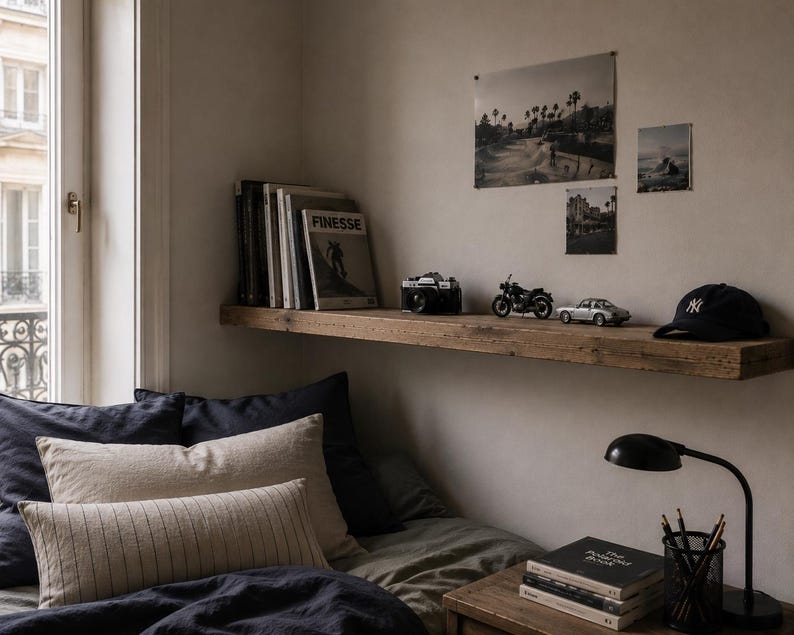 May include: A bedroom scene with a wooden shelf displaying a camera, motorcycle, car, and a baseball cap. The shelf also holds books and a magazine with the word "FINESSE". Below, a bed is made with dark blue and beige pillows and bedding.