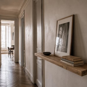 May include: A hallway with a wooden floor and white walls. A wooden shelf holds a framed black and white photograph, a small bowl, and several books. The hallway leads to a room with a window.