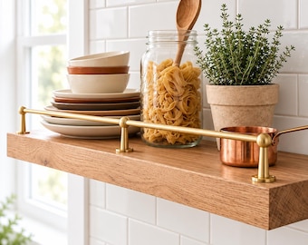 May include: A wooden wall shelf with a brass rail holds a stack of ceramic plates and bowls, a glass jar filled with pasta and a wooden spoon, a potted green herb, and a small copper pot against a white subway tile backsplash.