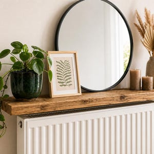 Può includere: Una mensola rustica in legno espone uno specchio rotondo con cornice nera, una stampa botanica incorniciata e una pianta in vaso con foglie verdi. Candele decorative e un vaso con erbe secche completano l'estetica naturale. La mensola è montata sopra un radiatore bianco.
