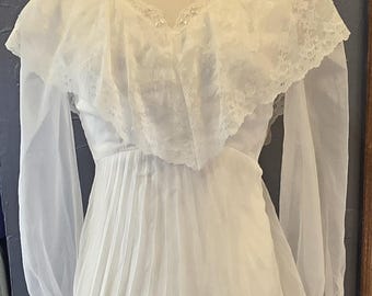 Puede incluir: Vestido de novia blanco vintage con cuello alto y mangas largas y transparentes. El corpiño presenta una superposición de encaje con delicados detalles de cuentas y una falda plisada. El vestido se exhibe en un maniquí.