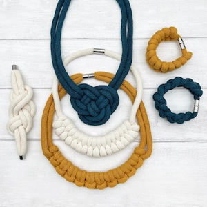 May include: A set of four macrame necklaces in teal, white, and mustard yellow. The necklaces are made of knotted rope and feature different styles and lengths.