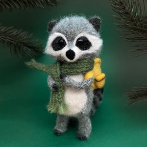 May include: A gray and white raccoon plush toy wearing a green scarf and holding a yellow banana. The raccoon has black eyes and a black nose.
