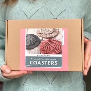 May include: A brown box with a pink label that says "Macrame Kit" and "Learn to make these stylish macrame Coasters". The label also has a picture of three macrame coasters in different colours: grey, beige, and pink.