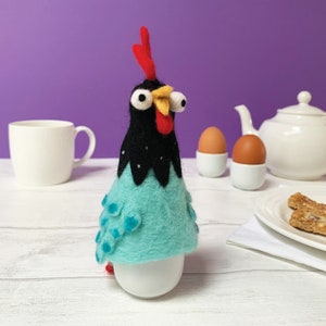 Puede incluir: Un calienta huevos de fieltro azul y negro con ojos saltones en forma de pollo. El pollo está sentado en un porta huevos blanco.